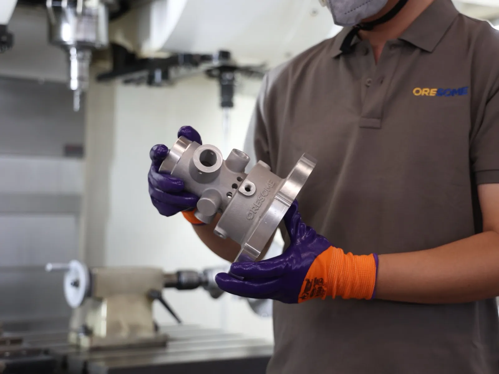 A machinist at a CNC mill holding a freshly machined pump head with ORESOME cast into the flange.